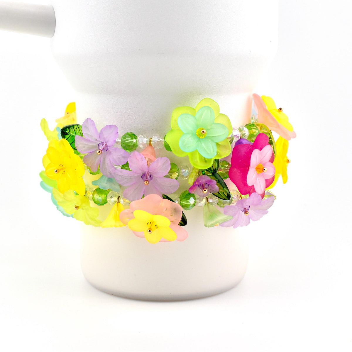 A colorful floral bracelet with yellow, pink, purple, and green flowers. The bracelet is adorned with clear beads and is wrapped around a white cylindrical object. The flowers have a translucent appearance.