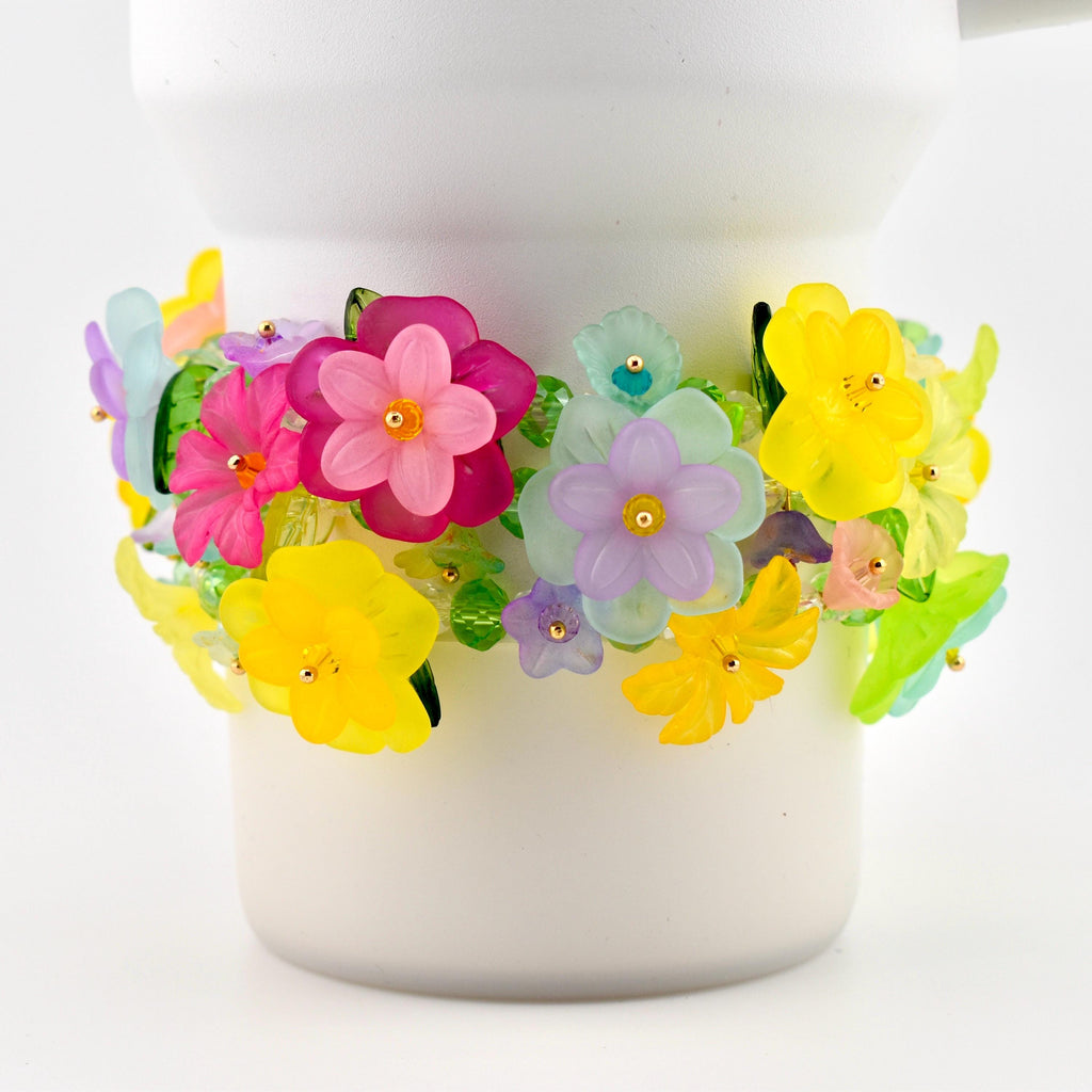 A colorful floral bracelet with yellow, pink, purple, and green flowers. The flowers are made of translucent plastic and are attached to a clear beaded band. The bracelet is wrapped around a white cylindrical object.