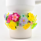 A white mug adorned with a vibrant floral bracelet. The bracelet features translucent flowers in shades of pink, yellow, purple, and green, with gold-colored centers. The flowers are arranged around the mug's body.