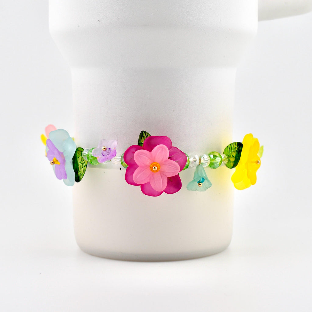 A floral bracelet with colorful flower beads. The bracelet features a large pink flower in the center, with smaller purple, blue, and yellow flowers. The bracelet is adorned with green leaves and clear beads.