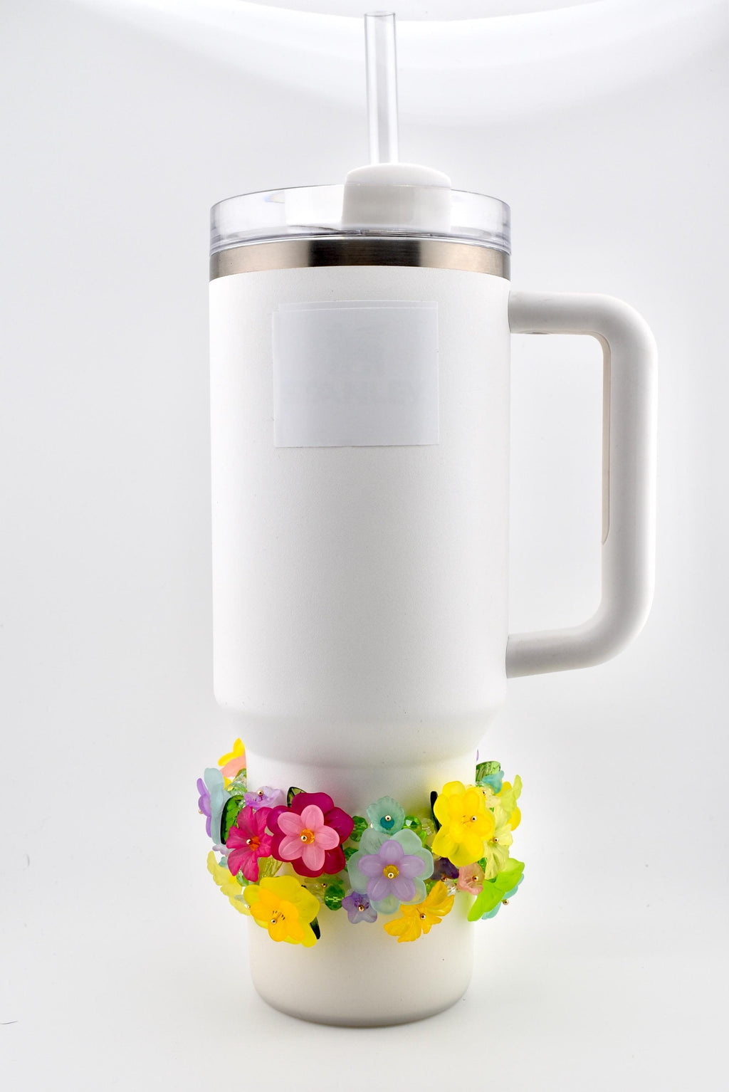 White insulated tumbler with a handle, clear lid, and straw. A decorative floral band in pink, yellow, purple, and green encircles the base. The tumbler has a white label with the brand name.