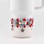 A stack of beaded bracelets in red, white, and blue, with star charms. The bracelets are wrapped around a white mug. The beads are various sizes and shapes, with some iridescent and pearl-like.