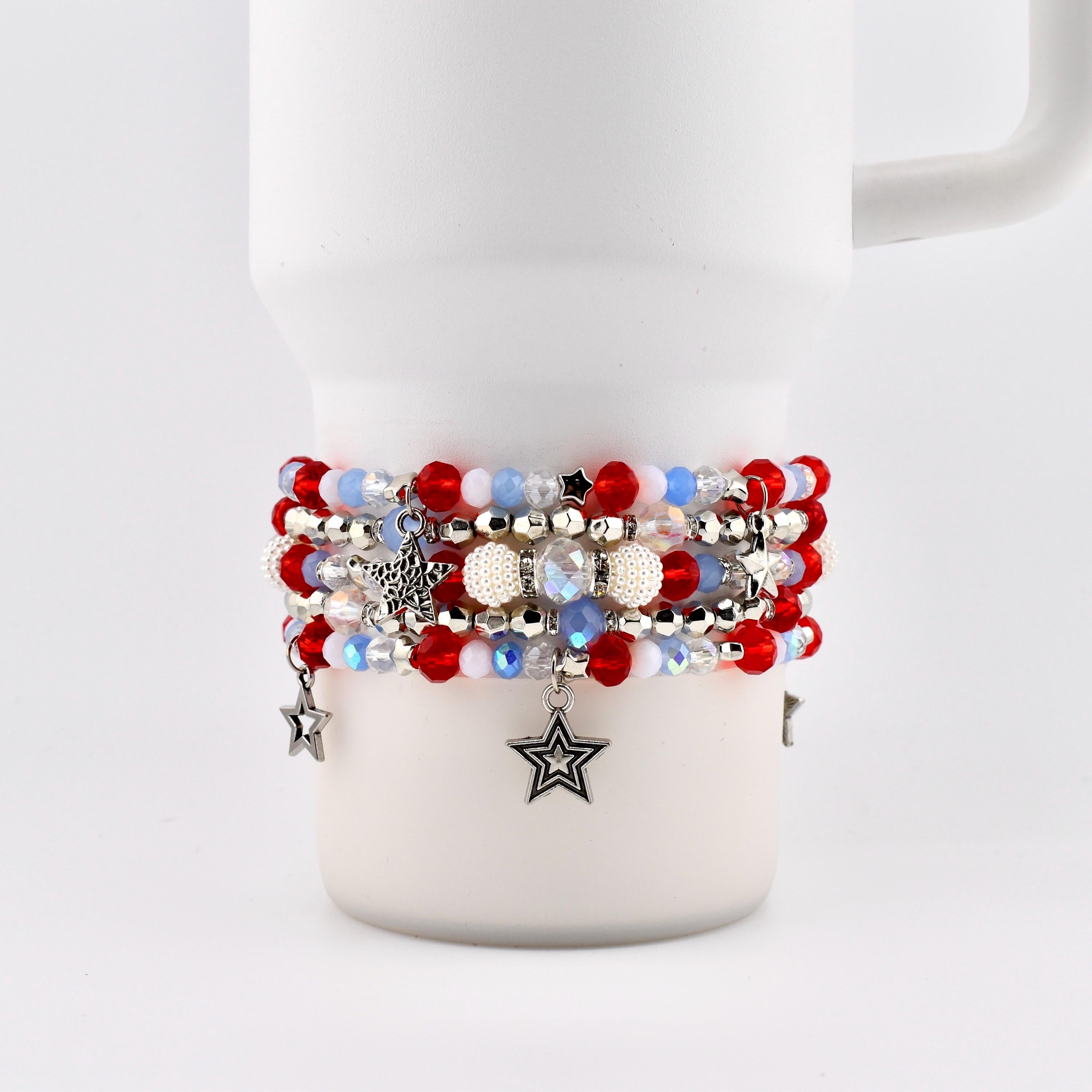 A stack of beaded bracelets in red, white, and blue, with star charms. The bracelets are wrapped around a white mug. The beads are various sizes and shapes, with some iridescent and pearl-like.