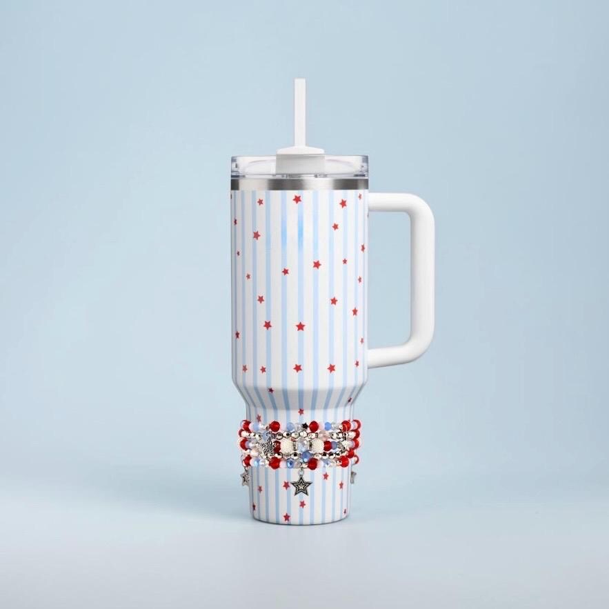 A white tumbler with a handle, a lid, and a straw. The tumbler has a pattern of blue vertical stripes and red stars. A decorative beaded bracelet with red, white, and blue beads and star charms is wrapped around the base.