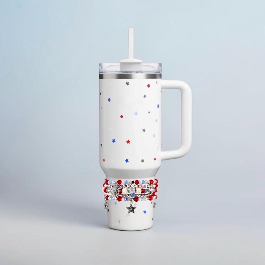White insulated tumbler with a handle, lid, and straw. The tumbler is decorated with red, white, and blue stars. A band of red, white, and blue beads and star charms adorns the bottom of the tumbler.