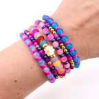 A collection of colorful beaded bracelets. The bracelets feature a variety of beads in shades of pink, blue, purple, and gold. Some bracelets include a white daisy-shaped bead. The bracelets are stacked on a wrist.