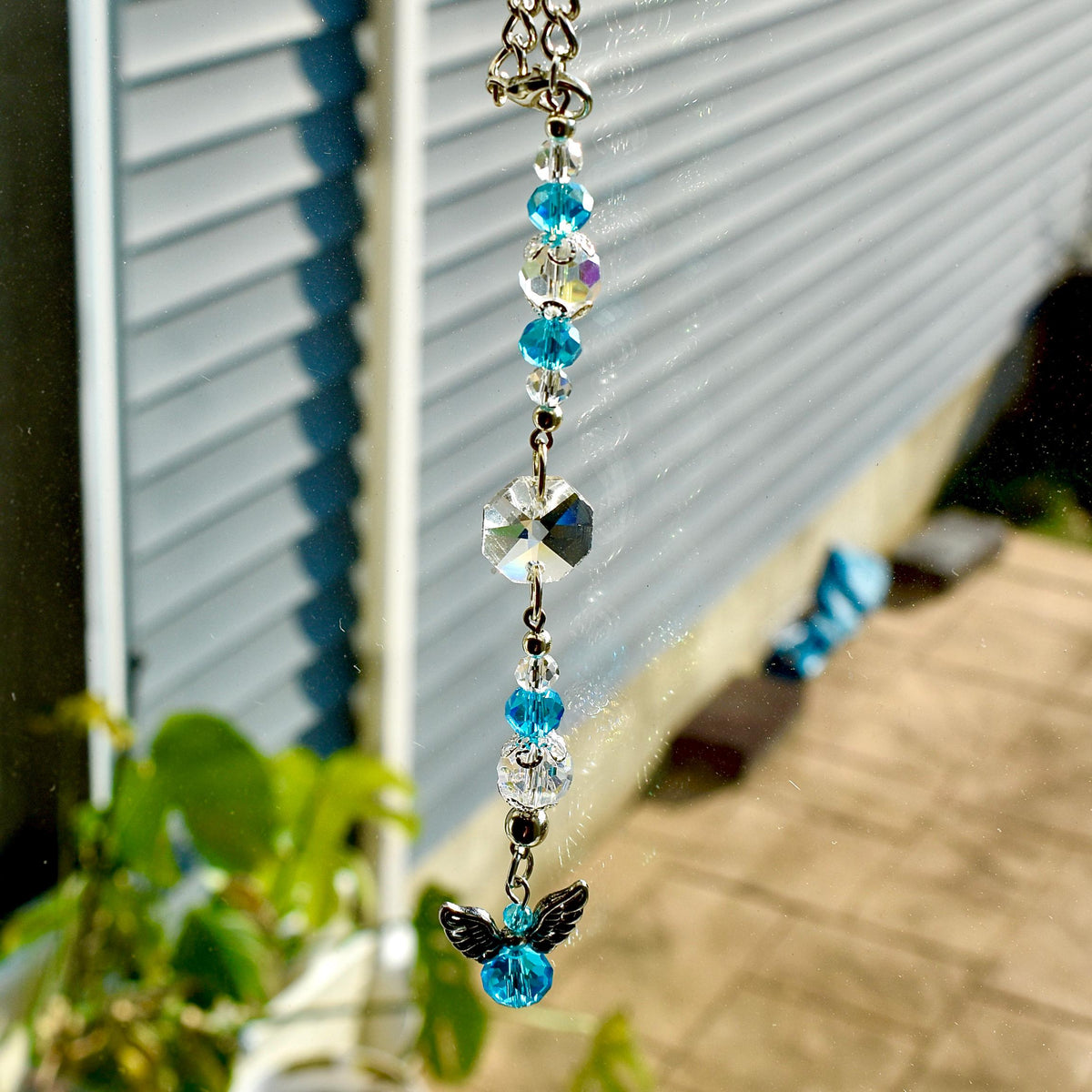 A decorative hanging ornament featuring a silver chain with clear and blue crystal beads. A large, faceted crystal and a small angel charm with blue crystal body and silver wings are at the bottom. The ornament is hanging in front of a window.