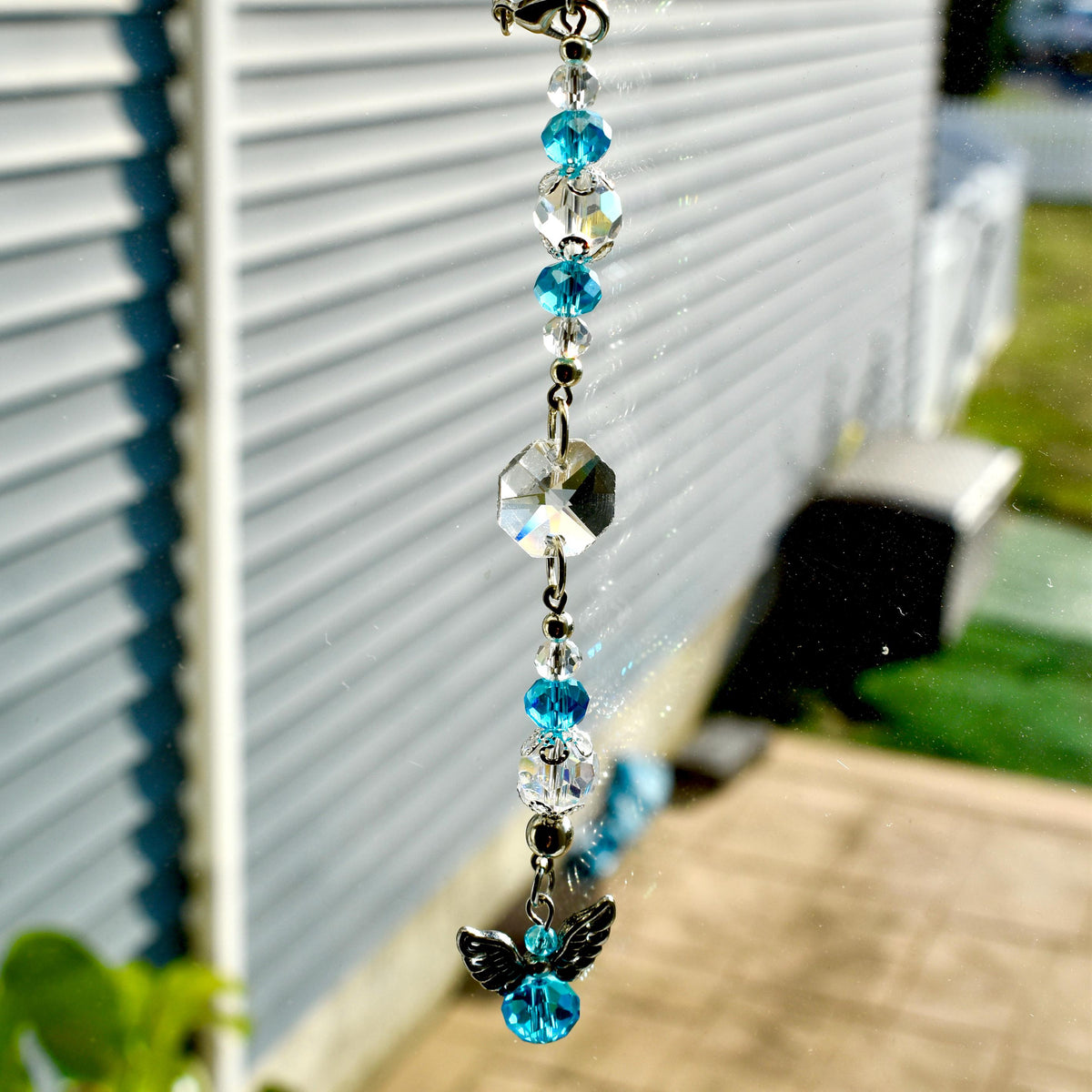 A decorative suncatcher featuring a series of clear and turquoise crystal beads and a faceted crystal pendant. The suncatcher is topped with a silver-colored clasp and has a small angel charm at the bottom.