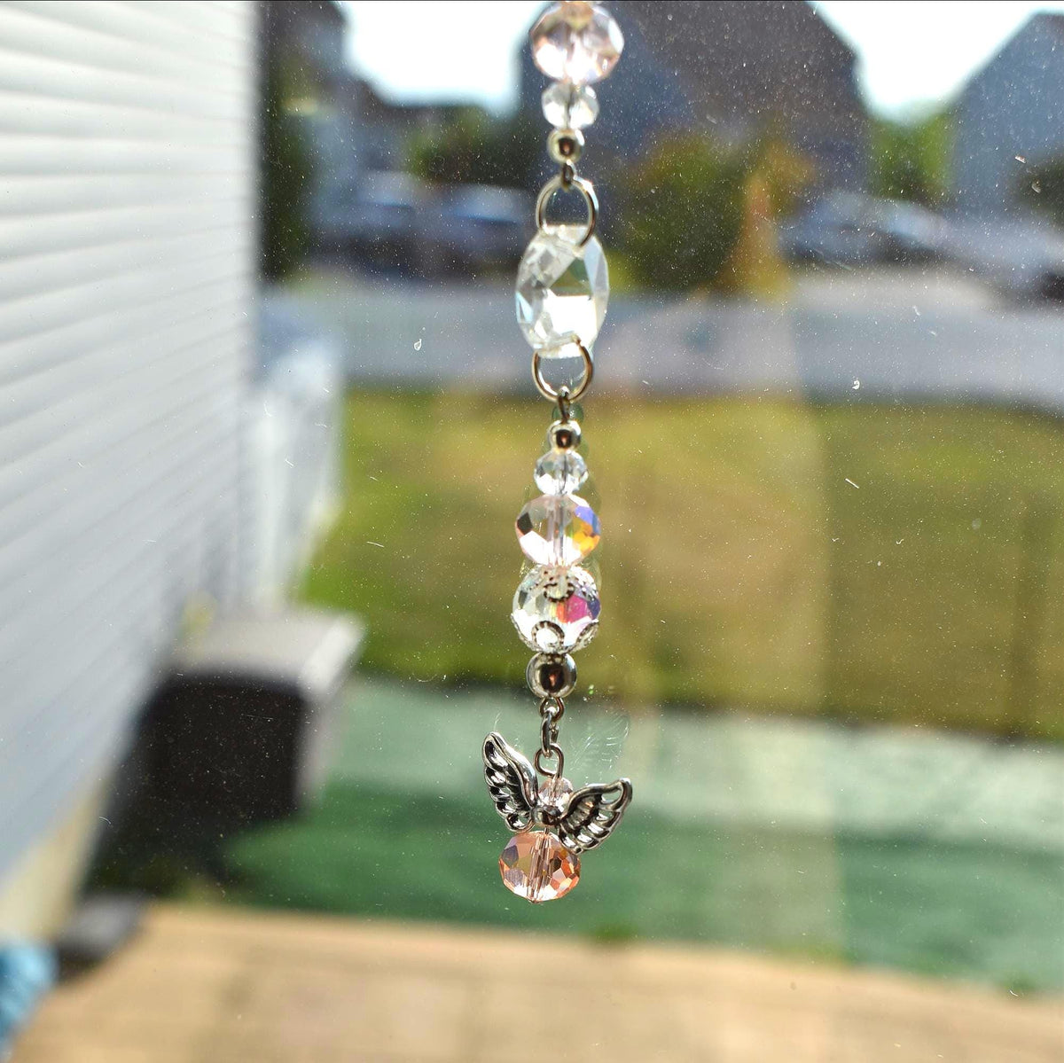 A decorative suncatcher with clear and iridescent crystal beads and a small angel charm. The angel charm has silver wings and a peach-colored crystal body. The suncatcher hangs from a silver chain.