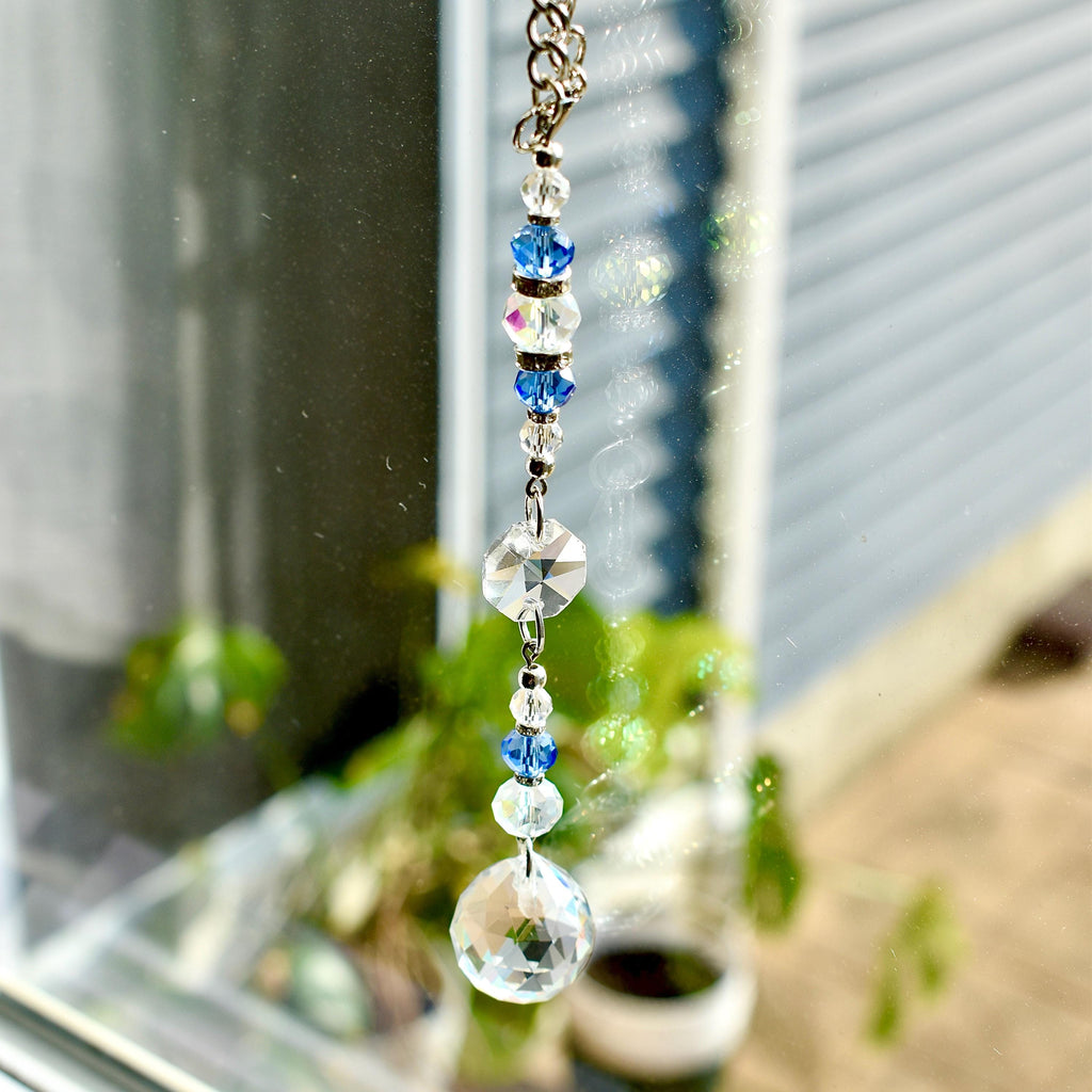 A decorative crystal suncatcher with a silver chain. The suncatcher features clear, blue, and iridescent crystal beads and a large faceted crystal ball at the bottom. The suncatcher is hanging in front of a window.