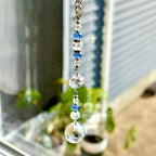 A decorative crystal suncatcher with a silver chain. The suncatcher features clear, blue, and iridescent crystal beads and a large faceted crystal ball at the bottom. The suncatcher is hanging in front of a window.