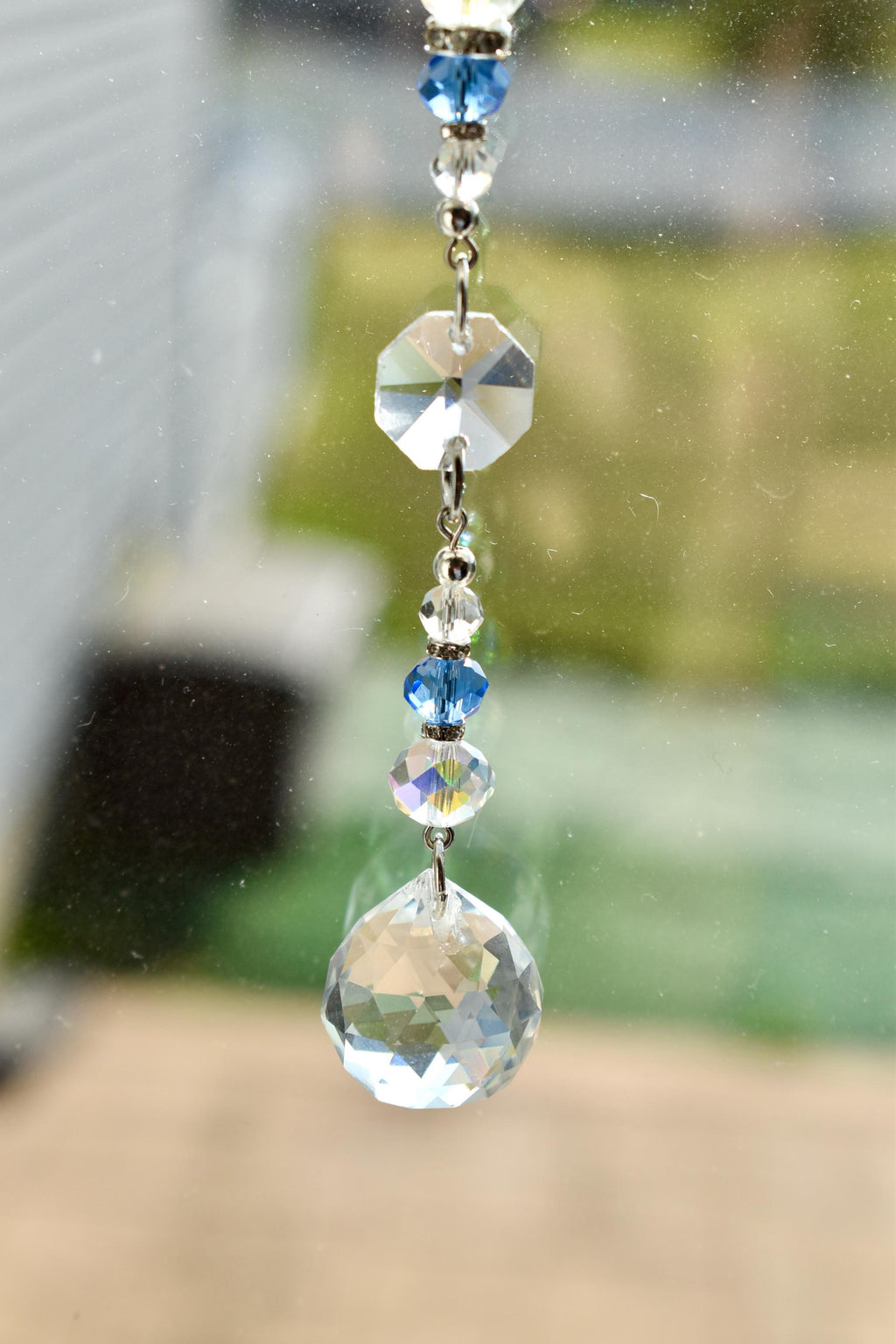 A decorative crystal suncatcher with a series of clear and blue crystal beads and a large, faceted crystal ball at the bottom. The suncatcher is suspended by a silver chain, reflecting light.