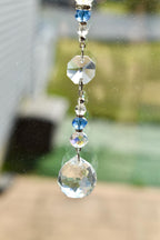A decorative crystal suncatcher with a series of clear and blue crystal beads and a large, faceted crystal ball at the bottom. The suncatcher is suspended by a silver chain, reflecting light.