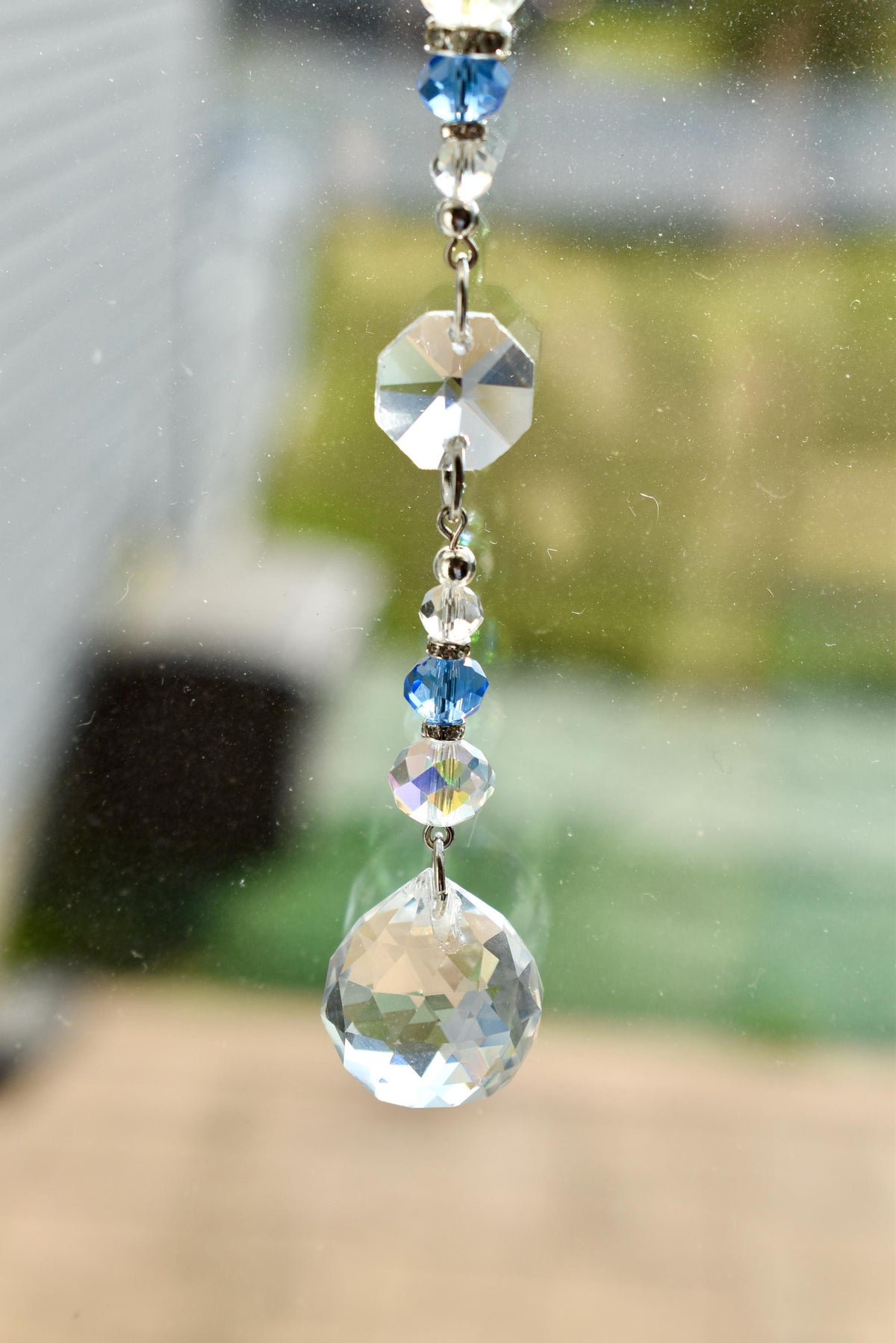 A decorative crystal suncatcher with a series of clear and blue crystal beads and a large, faceted crystal ball at the bottom. The suncatcher is suspended by a silver chain, reflecting light.