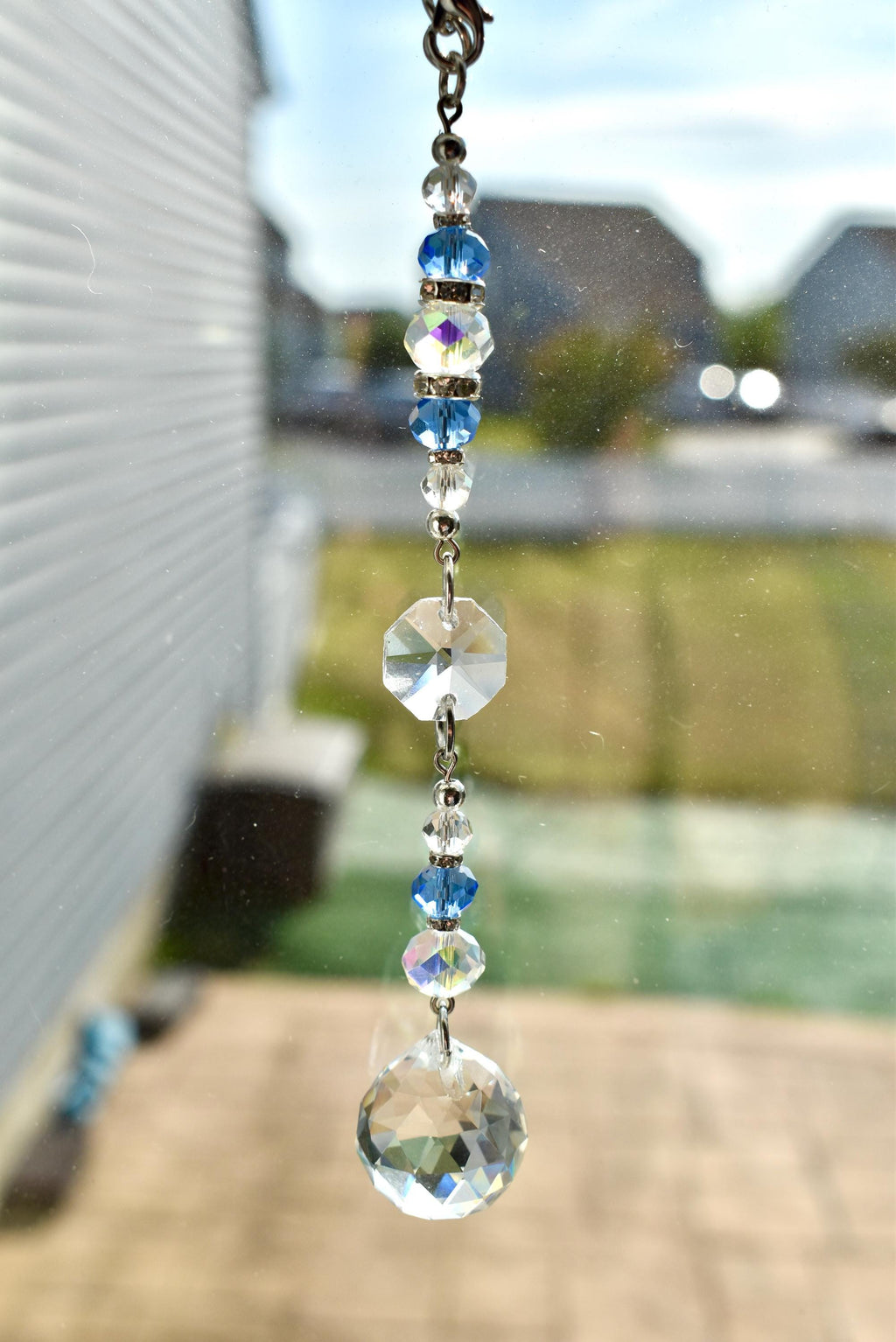 A decorative crystal suncatcher with clear, blue, and iridescent beads. The suncatcher features a faceted crystal ball and an octagonal crystal pendant, suspended by a silver chain. The beads are various sizes and shapes.