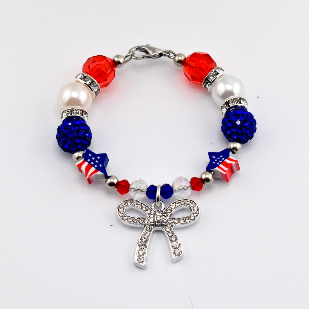 A beaded bracelet featuring red, white, and blue beads, including faceted red beads, white pearls, blue glitter spheres, and star-shaped beads with American flag patterns. A silver-toned bow charm encrusted with clear crystals hangs.