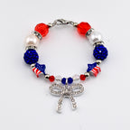 A beaded bracelet featuring red, white, and blue beads, including faceted red beads, white pearls, blue glitter spheres, and star-shaped beads with American flag patterns. A silver-toned bow charm encrusted with clear crystals hangs.
