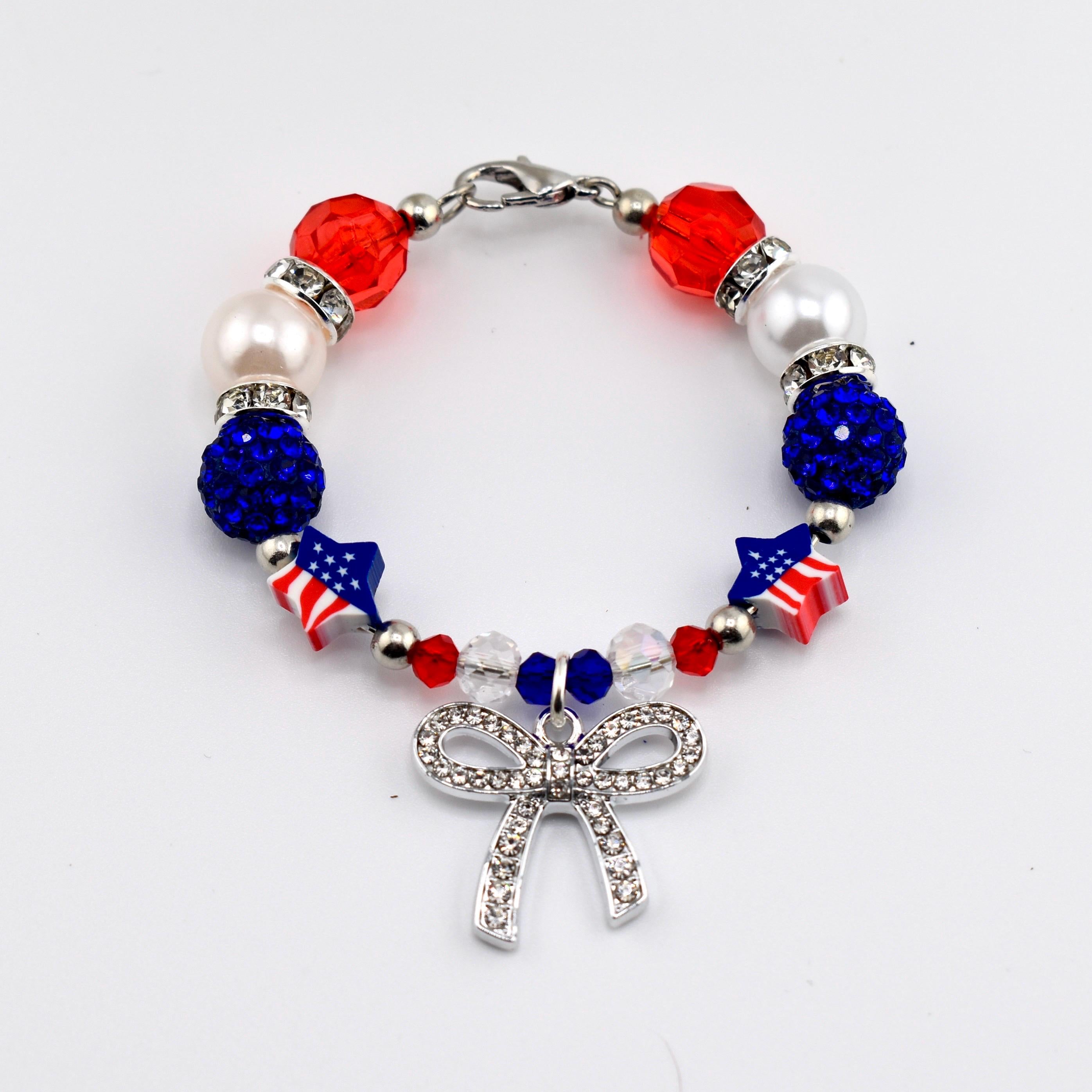 A beaded bracelet featuring red, white, and blue beads, including faceted red beads, white pearls, blue glitter spheres, and star-shaped beads with American flag patterns. A silver-toned bow charm encrusted with clear crystals hangs.