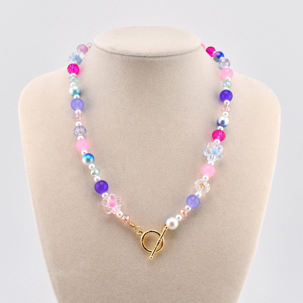 A colorful beaded necklace with pink, purple, blue, and white beads. The necklace features a gold-colored toggle clasp and is displayed on a mannequin. The beads are various sizes and shapes, creating a playful and vibrant accessory.