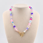 A colorful beaded necklace with pink, purple, blue, and white beads. The necklace features a gold-colored toggle clasp and is displayed on a mannequin. The beads are various sizes and shapes, creating a playful and vibrant accessory.