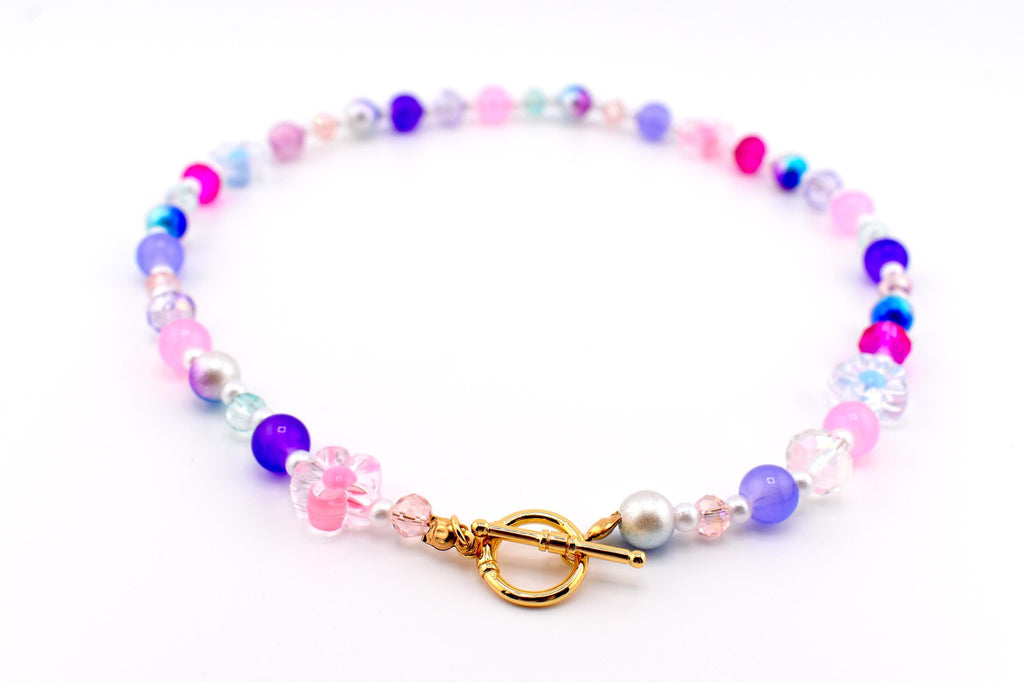 A colorful beaded necklace featuring a variety of beads in shades of pink, purple, blue, and clear. The necklace includes a gold-colored toggle clasp and a flower-shaped bead. The beads are strung together to create a delicate and eye-catching accessory.