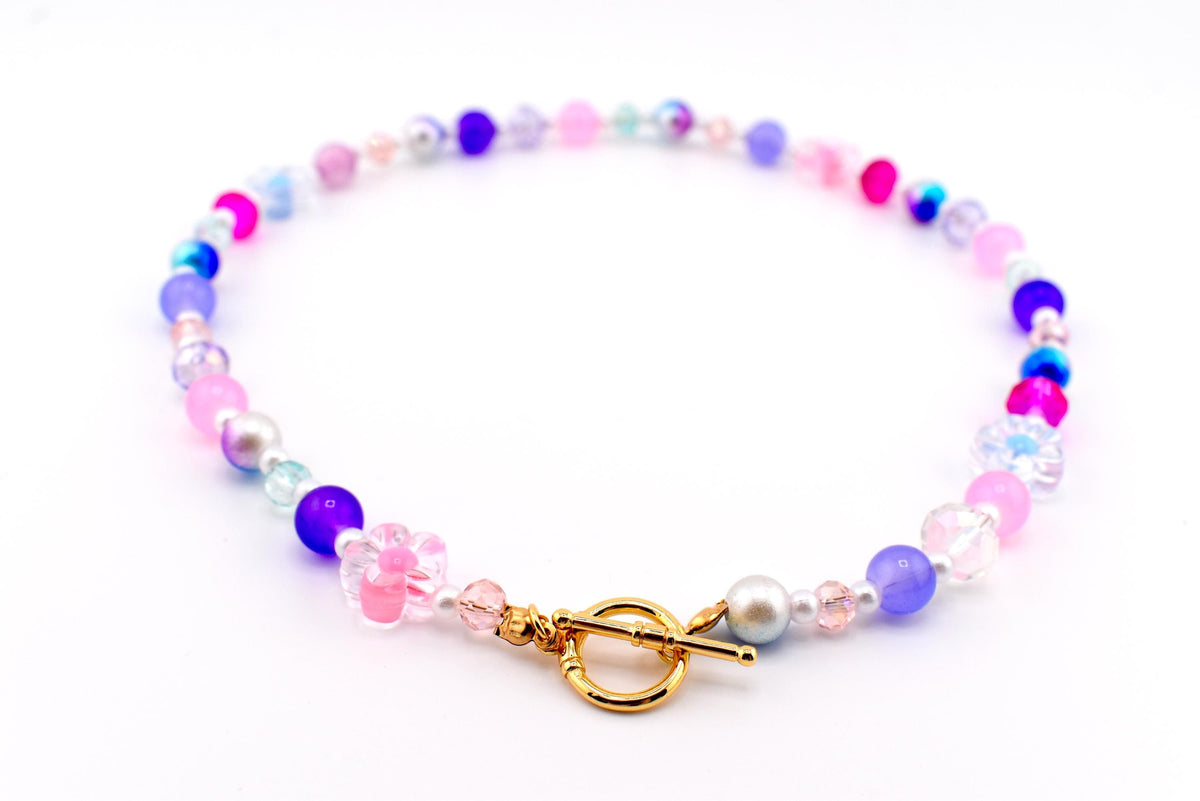 A colorful beaded necklace featuring a variety of beads in shades of pink, purple, blue, and clear. The necklace includes a gold-colored toggle clasp and a flower-shaped bead. The beads are strung together to create a delicate and eye-catching accessory.