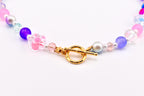 A colorful beaded necklace featuring a gold-tone toggle clasp. The necklace includes pink, purple, and iridescent beads, along with a clear flower-shaped bead with pink accents. The beads are strung together with a delicate chain.