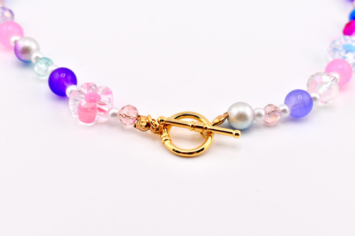 A colorful beaded necklace featuring a gold-tone toggle clasp. The necklace includes pink, purple, and iridescent beads, along with a clear flower-shaped bead with pink accents. The beads are strung together with a delicate chain.