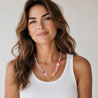 A colorful beaded necklace with pink, purple, and white beads, and a gold-colored clasp. The necklace is worn by a person wearing a white tank top.