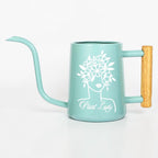 A teal watering can with a long, curved spout and a wooden handle. The can features a white graphic of a woman's face with a floral crown and the words "Plant Lady".