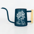 A dark blue watering can with a long, curved spout and a wooden handle. The can features a white graphic of a woman's face with a floral crown and the text "Plant Lady".