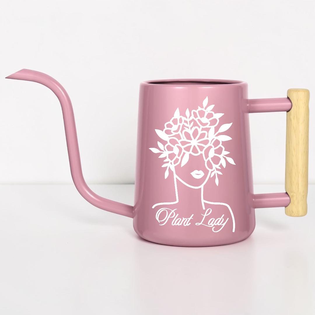 A pink metal watering can with a long spout and wooden handle. The can features a white graphic of a woman's face with a floral crown and the text "Plant Lady".