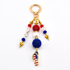 A patriotic keychain with a gold ring and dangling charms. The charms include red, white, and blue beads, gold stars, and a flip-flop charm with the American flag design. The word "USA" is spelled out with white letter beads.