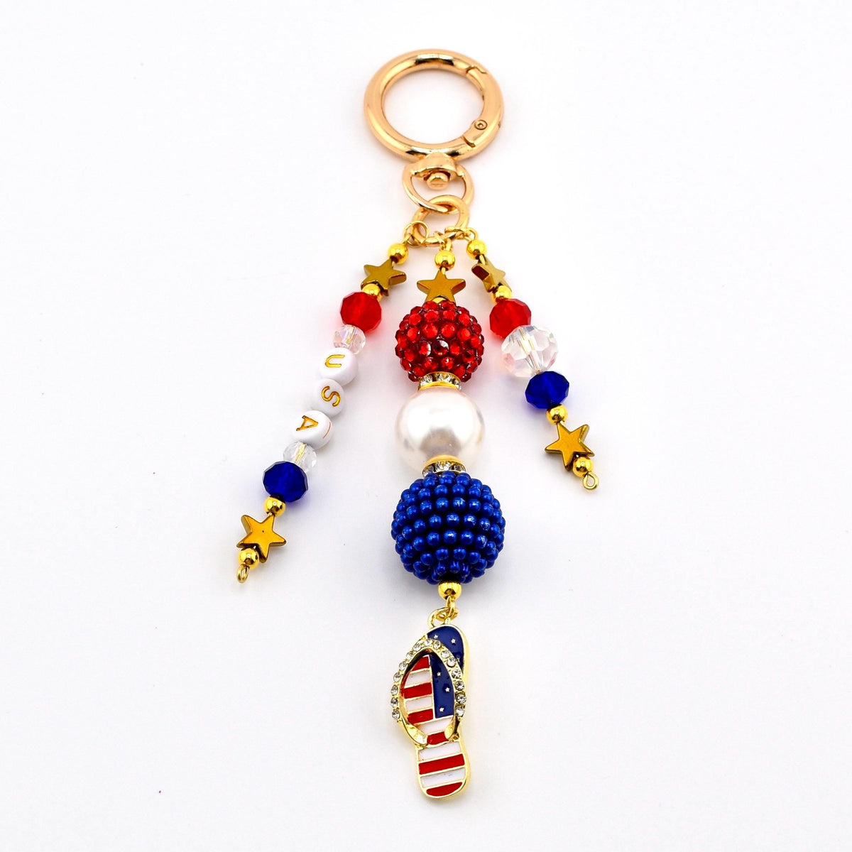 A patriotic keychain with a gold ring and dangling charms. The charms include red, white, and blue beads, gold stars, and a flip-flop charm with the American flag design. The word "USA" is spelled out with white letter beads.