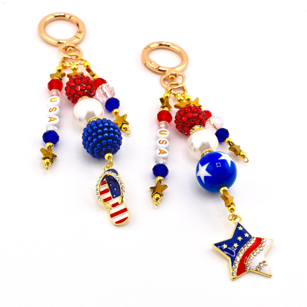 Two keychains with patriotic themes. Each keychain features gold-colored hardware, red, white, and blue beads, and star charms. One has a flip-flop charm with the American flag, and the other has a star charm with the American flag.