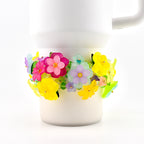 A white ceramic mug adorned with a colorful floral embellishment. The flowers are in shades of pink, yellow, purple, and green, with a clear, translucent appearance. The mug has a simple, classic design.