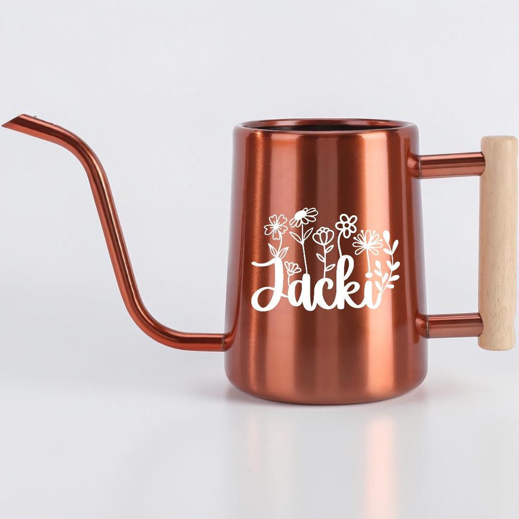 A copper-colored metal watering can with a long, slender spout and a light-colored wooden handle. The side of the watering can features a white graphic of various wildflowers and the name Jacki written in a cursive script font.