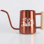 A copper-colored metal watering can with a long, slender spout and a light-colored wooden handle. The side of the watering can features a white graphic of various wildflowers and the name Jacki written in a cursive script font.