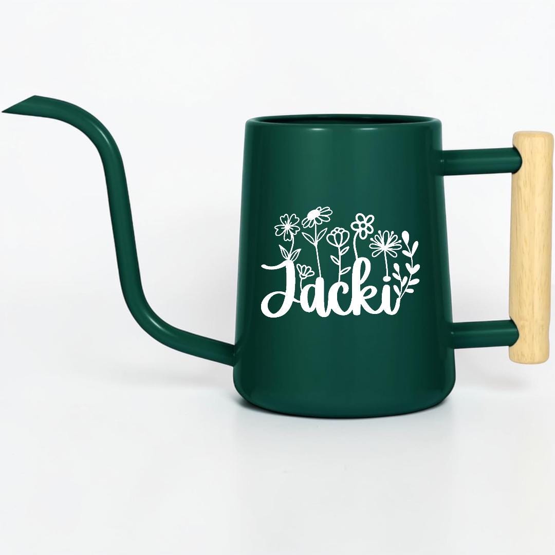 A dark green metal watering can with a long, slender spout and a light-colored wooden handle. The side of the watering can features a white graphic of various line-art flowers above the name Jacki written in a cursive script font.