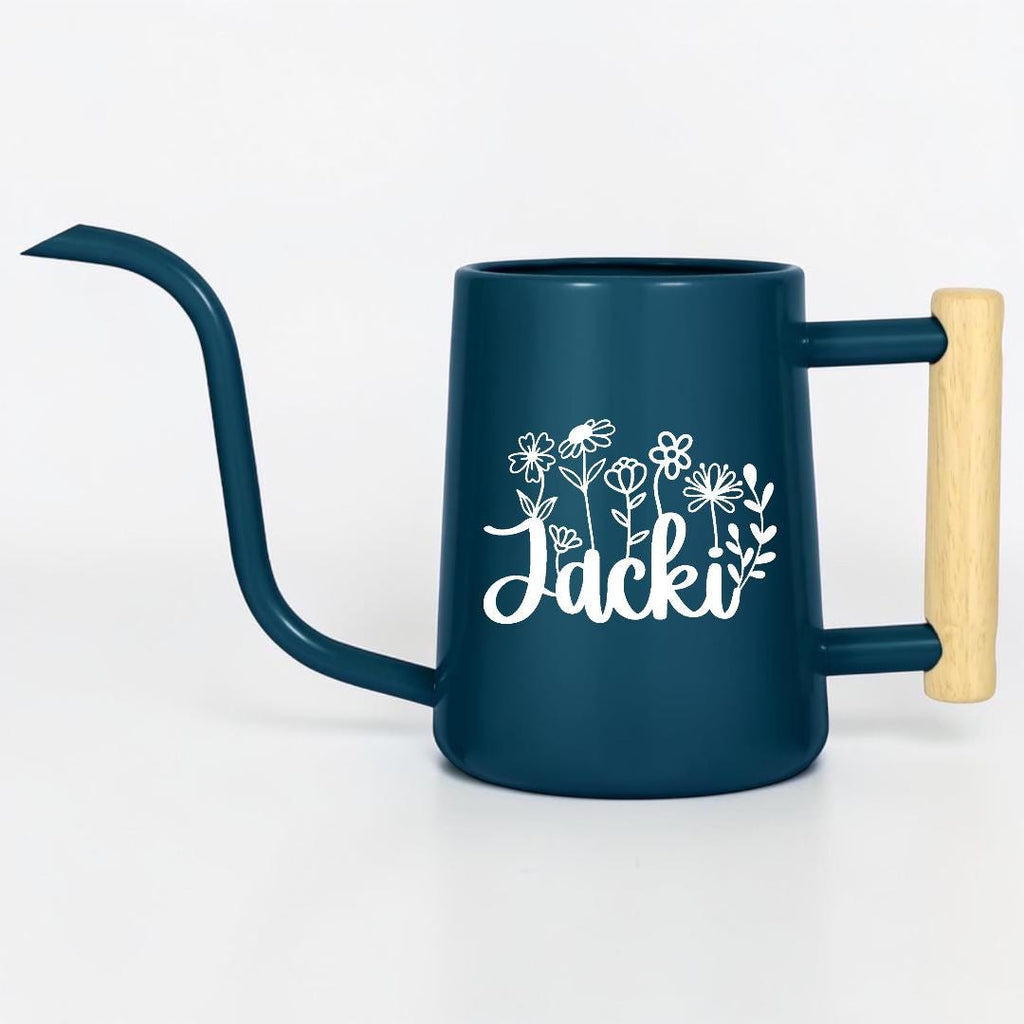 A dark blue metal watering can with a long, slender spout and a light-colored wooden handle. The side of the watering can features a white graphic design of various wildflowers and the name Jacki written in a cursive script font.