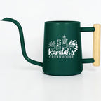 A dark green metal watering can with a long, slender spout and a light-colored wooden handle. The side of the can features a white graphic of four potted plants above the text Kamilah's GREENHOUSE in a mix of script and block fonts.