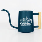 A dark blue metal watering can with a long, slender spout and a light-colored wooden handle. The side features a white graphic of four potted plants above the text "Kamilah's GREENHOUSE" in a mix of script and block lettering.