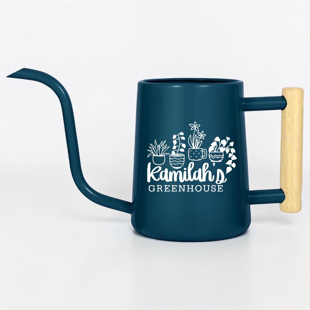 A dark blue metal watering can with a long, slender spout and a light-colored wooden handle. The side features a white graphic of four potted plants above the text "Kamilah's GREENHOUSE" in a mix of script and block lettering.