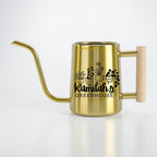 A gold-colored stainless steel watering can with a long, slender spout and a light-colored wooden handle. The side of the can features a black graphic of four potted plants and the text Kamilah's GREENHOUSE printed in black.
