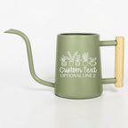 A sage green metal watering can with a long, slender spout and a light-colored wooden handle. The side features a white line art graphic of five potted plants above the text Custom Text and OPTIONAL LINE 2 in a white script font.