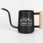 A matte black watering can with a long, slender spout and a light-colored wooden handle. The side of the can features a white line-art graphic of five potted plants above the text Custom Text and OPTIONAL LINE 2 in white font.