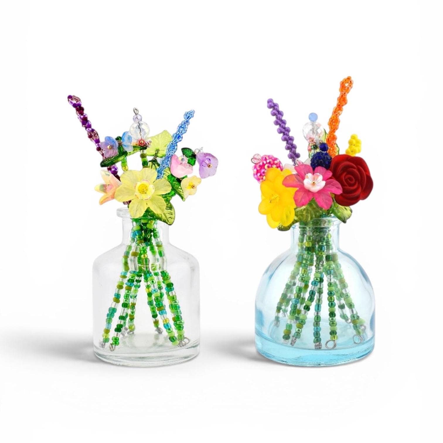 Two clear glass bottles containing artificial floral arrangements made from colorful beads. Each bouquet features various flower shapes in yellow, red, pink, and purple, with green beaded stems extending into the bottles.