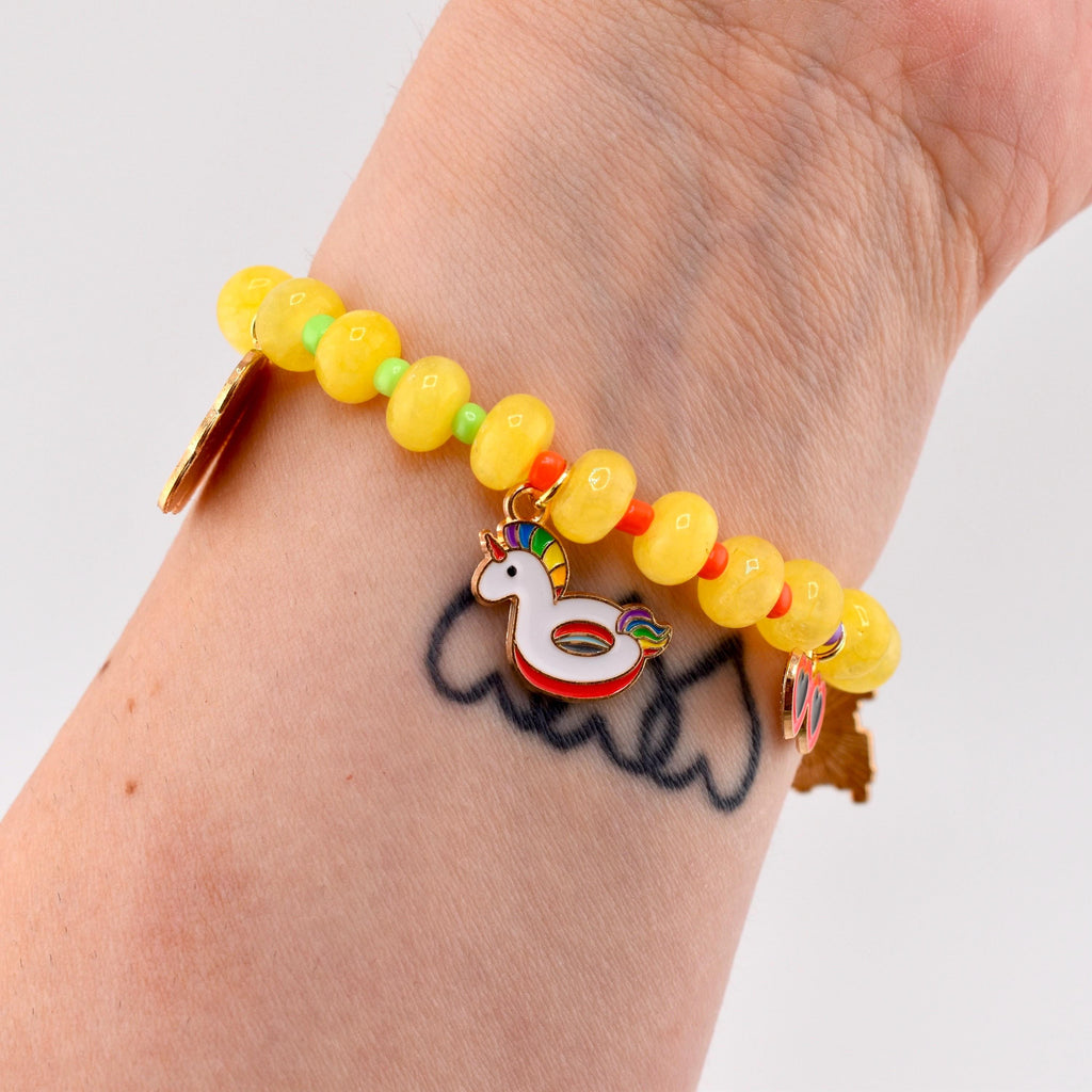 A beaded bracelet featuring translucent yellow circular beads separated by small green and orange spacers. The bracelet includes a gold-toned metal charm shaped like a white unicorn pool float with a rainbow mane and tail.