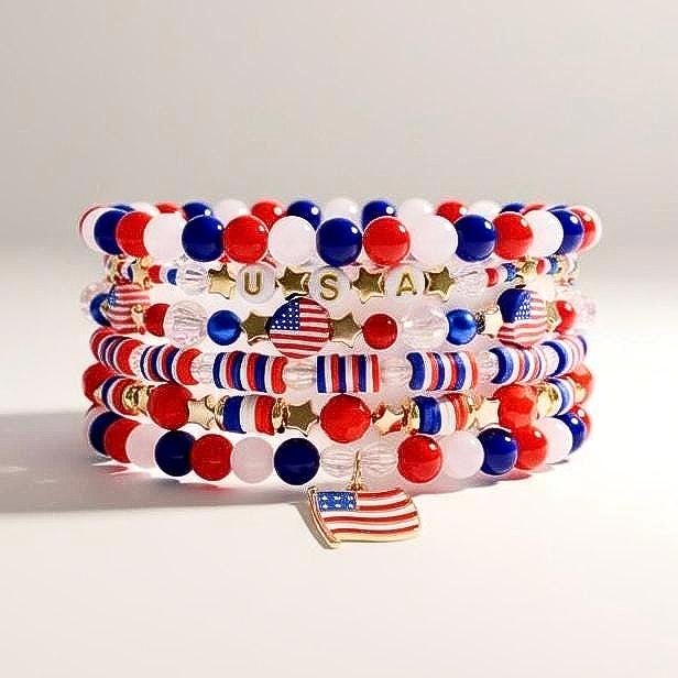 Stack of beaded bracelets in red, white, and blue, with gold accents. Some beads spell "USA" and feature American flags. A gold charm of the American flag hangs from the bottom bracelet. Patriotic jewelry.