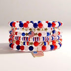 Stack of beaded bracelets in red, white, and blue, with gold accents. Some beads spell "USA" and feature American flags. A gold charm of the American flag hangs from the bottom bracelet. Patriotic jewelry.